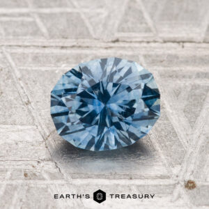 A faceted oval blue 1.32-carat heated Montana sapphire rests on a textured silver surface, with "Earth's Treasury" at the bottom.