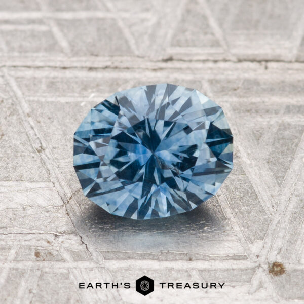 1.32-Carat Montana Sapphire (Heated) A faceted oval blue 1.32-carat heated Montana sapphire rests on a textured silver surface, with "Earth's Treasury" at the bottom.