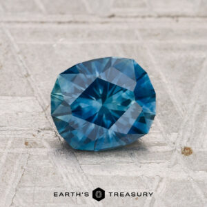 A 1.17-carat heated Montana sapphire rests on a textured gray surface, with the "Earth's Treasury" logo and text at the bottom.