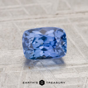 A 1.43-carat cushion-cut Ceylon Sapphire is displayed on a light-colored textured surface with the "Earth's Treasury" logo beneath it.