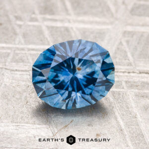 A 1.78-carat heated Montana sapphire with a visible inclusion sits on a textured metallic surface labeled "EARTH'S TREASURY.