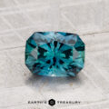 A faceted blue-green 1.58-carat Montana Sapphire (Heated) with an octagonal shape is shown on a textured gray surface. "EARTH'S TREASURY" appears at the bottom.