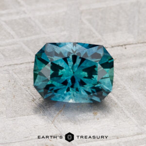 A faceted blue-green 1.58-carat Montana Sapphire (Heated) with an octagonal shape is shown on a textured gray surface. "EARTH'S TREASURY" appears at the bottom.
