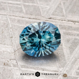A 1.31-carat heated Montana sapphire with a radiant cut rests on a textured gray surface, with the Earth's Treasury logo displayed at the bottom.