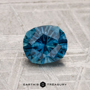 A 1.14-carat heated Montana sapphire sits on a textured gray surface, with "EARTH'S TREASURY" and a hexagon logo displayed beneath it.