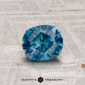 A 1.12-carat heated Montana sapphire with a geometric cut is shown on a textured gray surface, featuring the "Earth's Treasury" text and logo at the bottom.