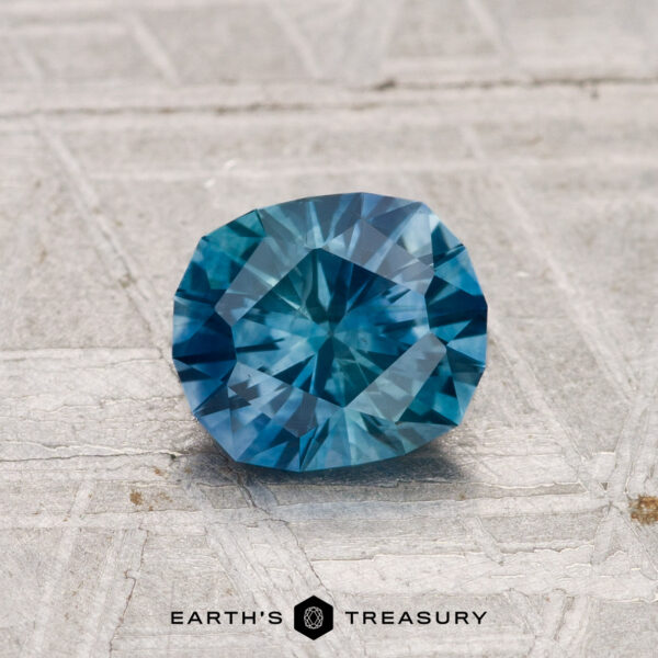 1.12-Carat Montana Sapphire (Heated) A 1.12-carat heated Montana sapphire with a geometric cut is shown on a textured gray surface, featuring the "Earth's Treasury" text and logo at the bottom.