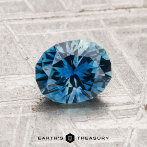 A 1.30-carat heated Montana sapphire is displayed on a textured gray surface, with the "EARTH'S TREASURY" text and logo at the bottom.