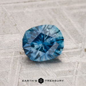 A 1.11-carat heated Montana sapphire with geometric facets rests on a textured gray surface. "EARTH'S TREASURY" is displayed at the bottom of the image.