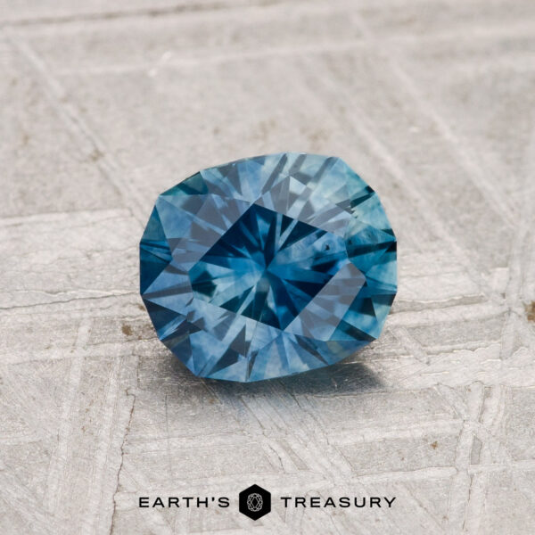 1.11-Carat Montana Sapphire (Heated) A 1.11-carat heated Montana sapphire with geometric facets rests on a textured gray surface. "EARTH'S TREASURY" is displayed at the bottom of the image.