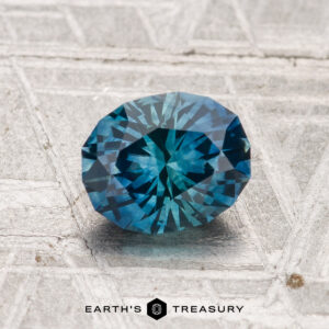 A 1.28-carat oval-cut blue-green Montana sapphire (heated) rests on a textured gray surface. "Earth's Treasury" logo and text are visible at the bottom of the image.