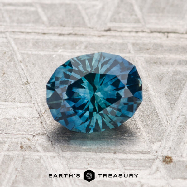 1.28-Carat Montana Sapphire (Heated) A 1.28-carat oval-cut blue-green Montana sapphire (heated) rests on a textured gray surface. "Earth's Treasury" logo and text are visible at the bottom of the image.