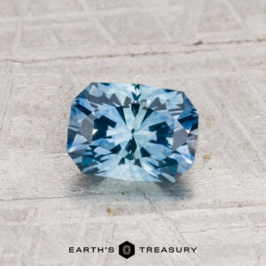 A 1.08-carat heated Montana sapphire, cut in a blue rectangular shape with intricate facets, is displayed on a light textured surface. The "Earth’s Treasury" logo is shown at the bottom.