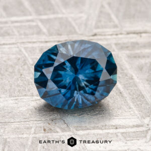 A 1.70-carat heated Montana sapphire with a deep blue facet is displayed on a textured surface, featuring the "Earth's Treasury" logo below.