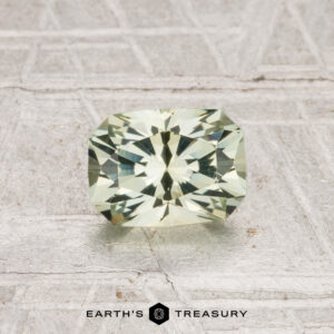 A rectangular, pale yellow-green 1.02-carat heated Montana sapphire rests on a textured gray surface, with "EARTH'S TREASURY" below.