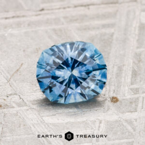 A 1.05-carat heated Montana sapphire with a geometric cut is showcased on a textured metallic surface featuring the "EARTH'S TREASURY" logo at the bottom.