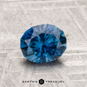 A 1.28-carat heated Montana sapphire gemstone is displayed on a textured light gray surface with "EARTH'S TREASURY" at the bottom.