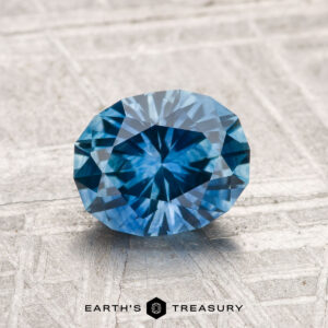 A 1.69-carat Montana Sapphire (heated), faceted in an oval shape, is displayed on a textured light gray surface with "EARTH'S TREASURY" at the bottom.