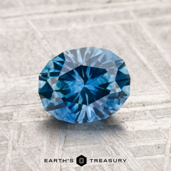 1.69-Carat Montana Sapphire (Heated) A 1.69-carat Montana Sapphire (heated), faceted in an oval shape, is displayed on a textured light gray surface with "EARTH'S TREASURY" at the bottom.