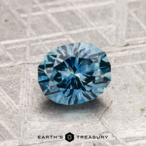 A 1.26-carat oval-cut heated Montana sapphire with intricate facets rests on a textured gray surface, featuring the "EARTH'S TREASURY" text and logo below.