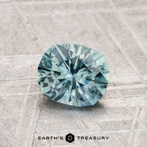 A 1.04-carat heated Montana sapphire, faceted in blue-green, rests on a light, textured surface with "EARTH'S TREASURY" at the bottom.