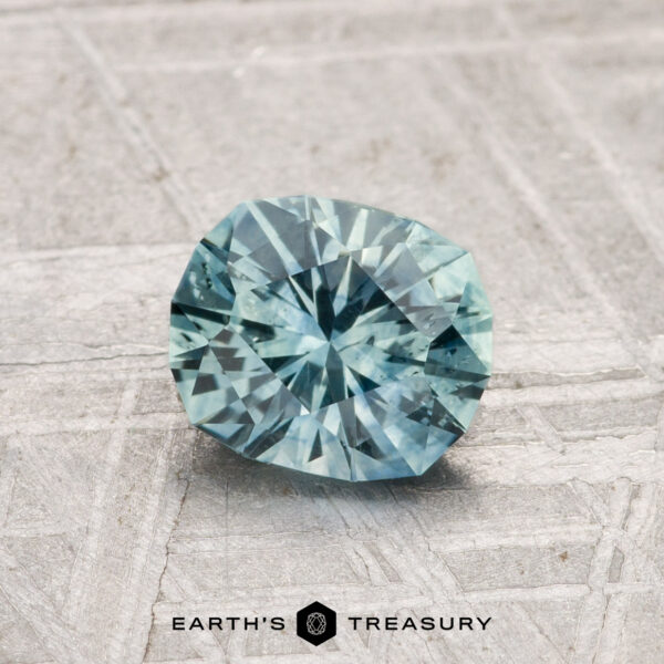 1.04-Carat Montana Sapphire (Heated) A 1.04-carat heated Montana sapphire, faceted in blue-green, rests on a light, textured surface with "EARTH'S TREASURY" at the bottom.