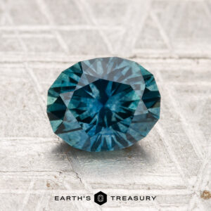 A 1.67-carat heated Montana sapphire, round and faceted, is shown on a pale, patterned surface with the Earth’s Treasury logo at the bottom.