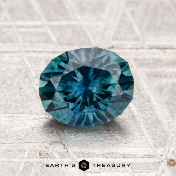 1.67-Carat Montana Sapphire (Heated) A 1.67-carat heated Montana sapphire, round and faceted, is shown on a pale, patterned surface with the Earth’s Treasury logo at the bottom.