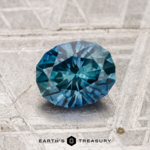 A 1.25-carat heated Montana sapphire with blue-green facets rests on a textured gray surface, with "EARTH'S TREASURY" visible at the bottom.
