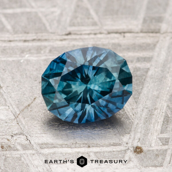 A 1.25-carat heated Montana sapphire with blue-green facets rests on a textured gray surface, with "EARTH'S TREASURY" visible at the bottom.