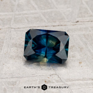 A rectangular, deep blue and teal 0.91-carat heated Montana sapphire rests on a textured gray surface with "EARTH'S TREASURY" below.
