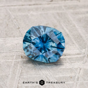 A 1.03-carat heated Montana sapphire gemstone is showcased on a textured gray surface with the "Earth's Treasury" logo visible at the bottom.
