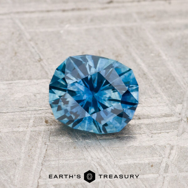 1.03-Carat Montana Sapphire (Heated) A 1.03-carat heated Montana sapphire gemstone is showcased on a textured gray surface with the "Earth's Treasury" logo visible at the bottom.