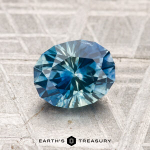 A faceted blue and green 1.64-carat heated Montana sapphire rests on a textured gray surface, with the text "EARTH'S TREASURY" at the bottom.