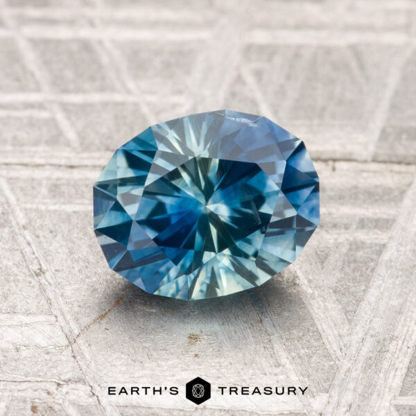 1.64-Carat Montana Sapphire (Heated) A faceted blue and green 1.64-carat heated Montana sapphire rests on a textured gray surface, with the text "EARTH'S TREASURY" at the bottom.