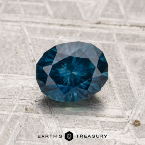 A 1.25-carat heated Montana sapphire with a deep blue hue rests on a textured gray surface, with "Earth's Treasury" displayed at the bottom.