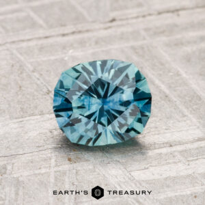 A 1.03-carat heated Montana sapphire with a blue square cushion cut rests on a textured gray surface, with "EARTH'S TREASURY" text below.