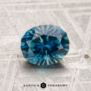 A 1.63-carat heated Montana sapphire, faceted in an oval shape and teal-blue in color, rests on a textured light surface with "EARTH'S TREASURY" below.