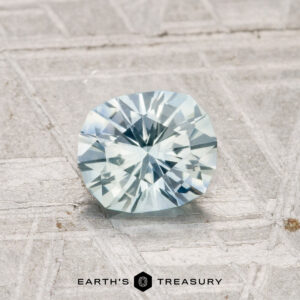 A 0.99-carat cushion-cut Montana Sapphire (heated), light blue in color, rests on a textured gray surface with "Earth's Treasury" displayed at the bottom.