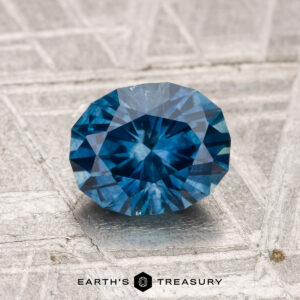A 1.62-carat heated Montana sapphire, cut in a faceted oval shape, is displayed on a textured gray surface with "EARTH'S TREASURY" and a hexagonal logo below.