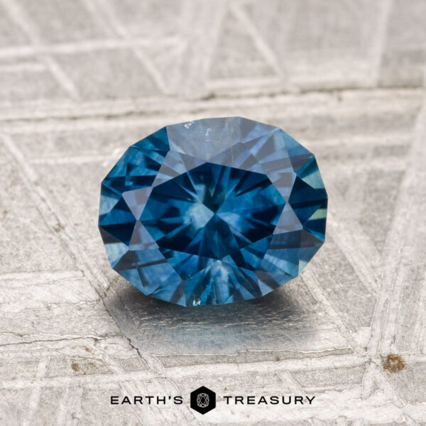1.62-Carat Montana Sapphire (Heated) A 1.62-carat heated Montana sapphire, cut in a faceted oval shape, is displayed on a textured gray surface with "EARTH'S TREASURY" and a hexagonal logo below.