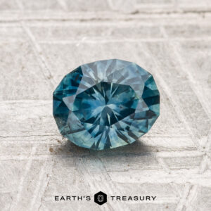 A 1.22-carat heated Montana sapphire, faceted in an oval cut and blue in color, rests on a textured, light surface featuring the "EARTH'S TREASURY" logo below.