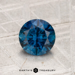 A 1.27-carat heated Montana sapphire with a round brilliant cut is displayed on a light textured surface, featuring the "Earth's Treasury" logo at the bottom.