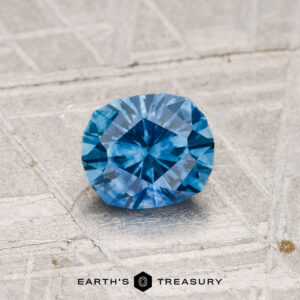 A faceted 0.88-carat heated Montana sapphire rests on a light-textured surface, with "EARTH'S TREASURY" and logo displayed at the bottom.