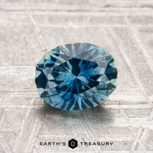 A 1.54-carat heated Montana Sapphire with geometric faceting is showcased on a textured surface, featuring the "EARTH'S TREASURY" logo at the bottom.