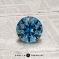 A round, faceted 0.96-carat heated Montana sapphire is shown on a light, textured surface with "EARTH'S TREASURY" displayed below it.