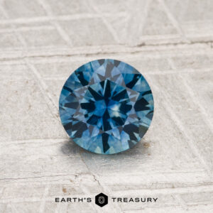 A round, faceted 0.96-carat heated Montana sapphire is shown on a light, textured surface with "EARTH'S TREASURY" displayed below it.