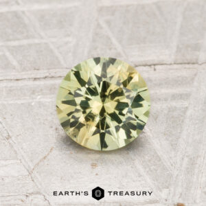 A 0.92-carat heated Montana sapphire, round and faceted with a pale yellow-green hue, is shown on a textured surface with "EARTH'S TREASURY" at the bottom.
