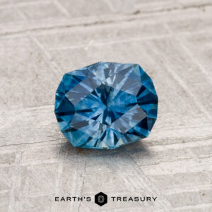 A 1.16-carat heated Montana sapphire, round and faceted, rests on a light textured surface with the Earth's Treasury logo displayed below.