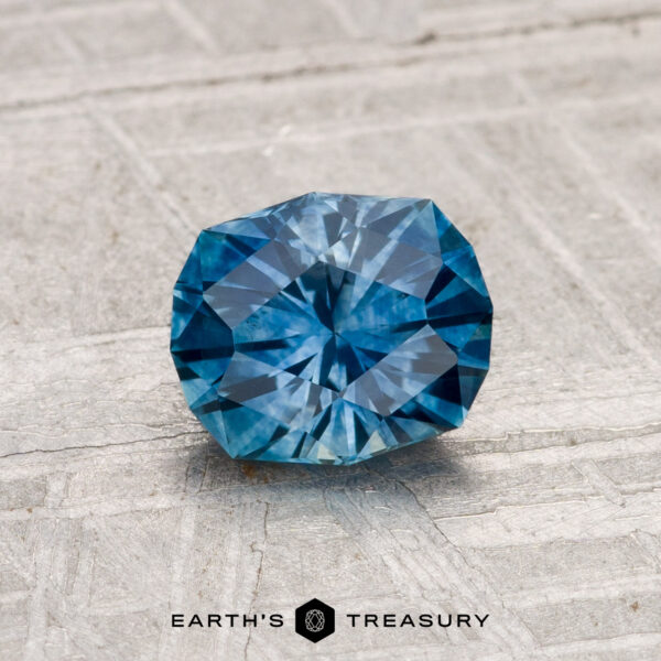 1.16-Carat Montana Sapphire (Heated) A 1.16-carat heated Montana sapphire, round and faceted, rests on a light textured surface with the Earth's Treasury logo displayed below.